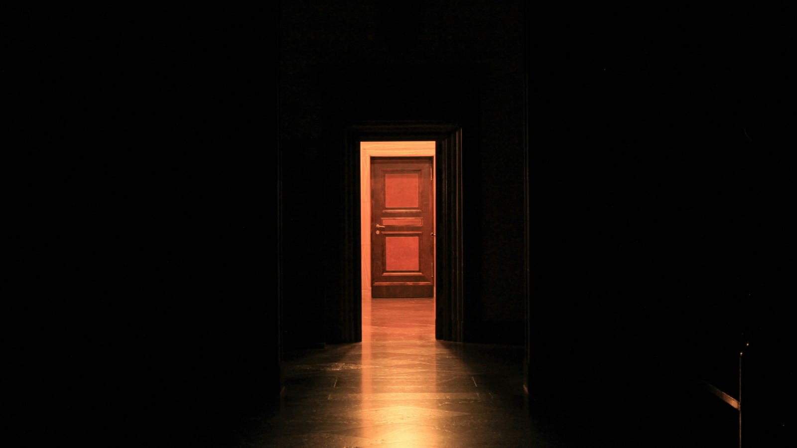 A dark corridor opens onto a warmly lit room, with a closed wooden door standing at the far end.